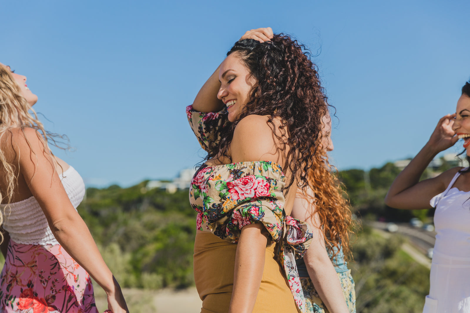 A picture of a women with very curly brown hair wearing a vibrant yellow floral top and yellow skirt she is smilling in the sunshine with one hand holding back her curls 